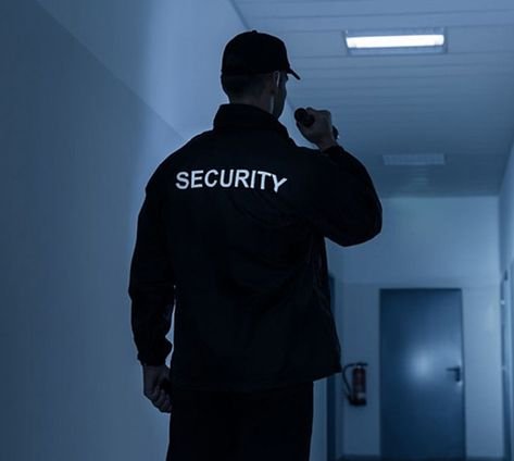 A security guard looking towards a closed door and using his flashlight to see in front of him.